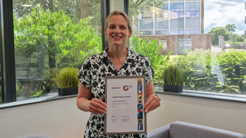 Person standing in an office setting with large windows and greenery outside, holding a framed certificate.